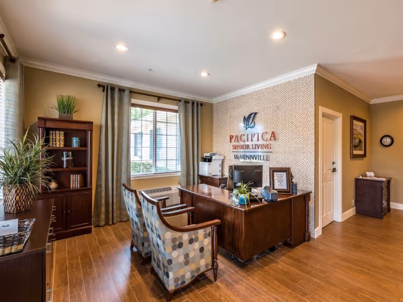 Reception area of Pacifica Senior Living McMinnville featuring a wooden desk with a computer, two patterned chairs, a bookshelf with decorative items, plants, and a window with curtains allowing natural light into the room.