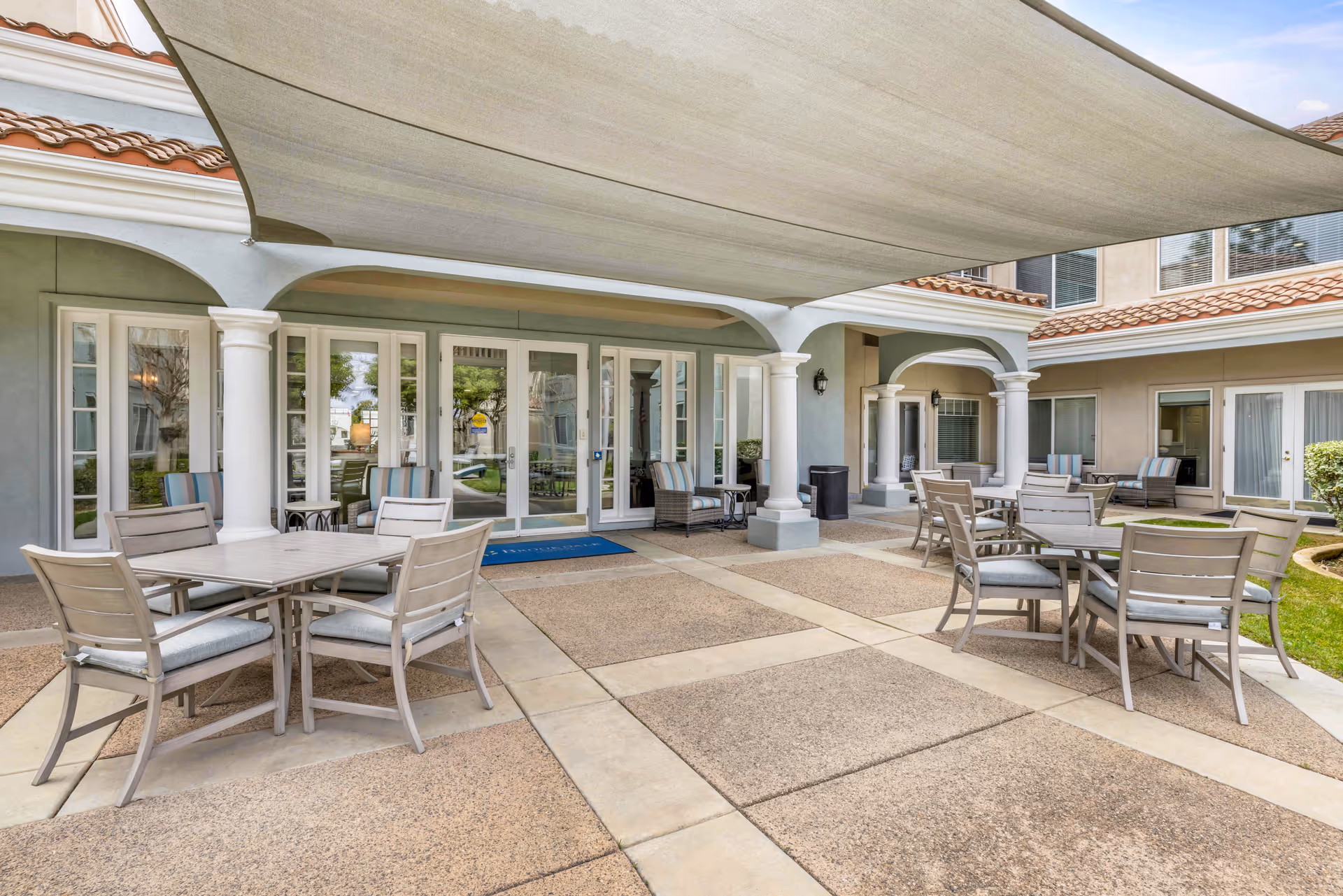 Outdoor patio area at Brookdale Murrieta with multiple tables and chairs arranged for seating. The space is shaded by a large canopy and surrounded by building walls with windows and doors leading inside. There are cushioned chairs and small side tables near the walls.