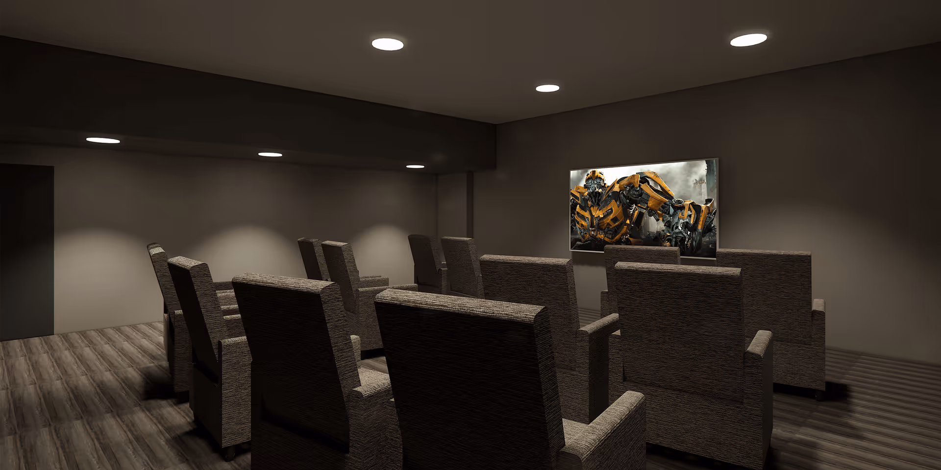 A small home theater room with two rows of cushioned armchairs facing a wall-mounted flat-screen TV displaying a scene from a Transformers movie. The room has dark walls, recessed ceiling lights, and wood flooring.
