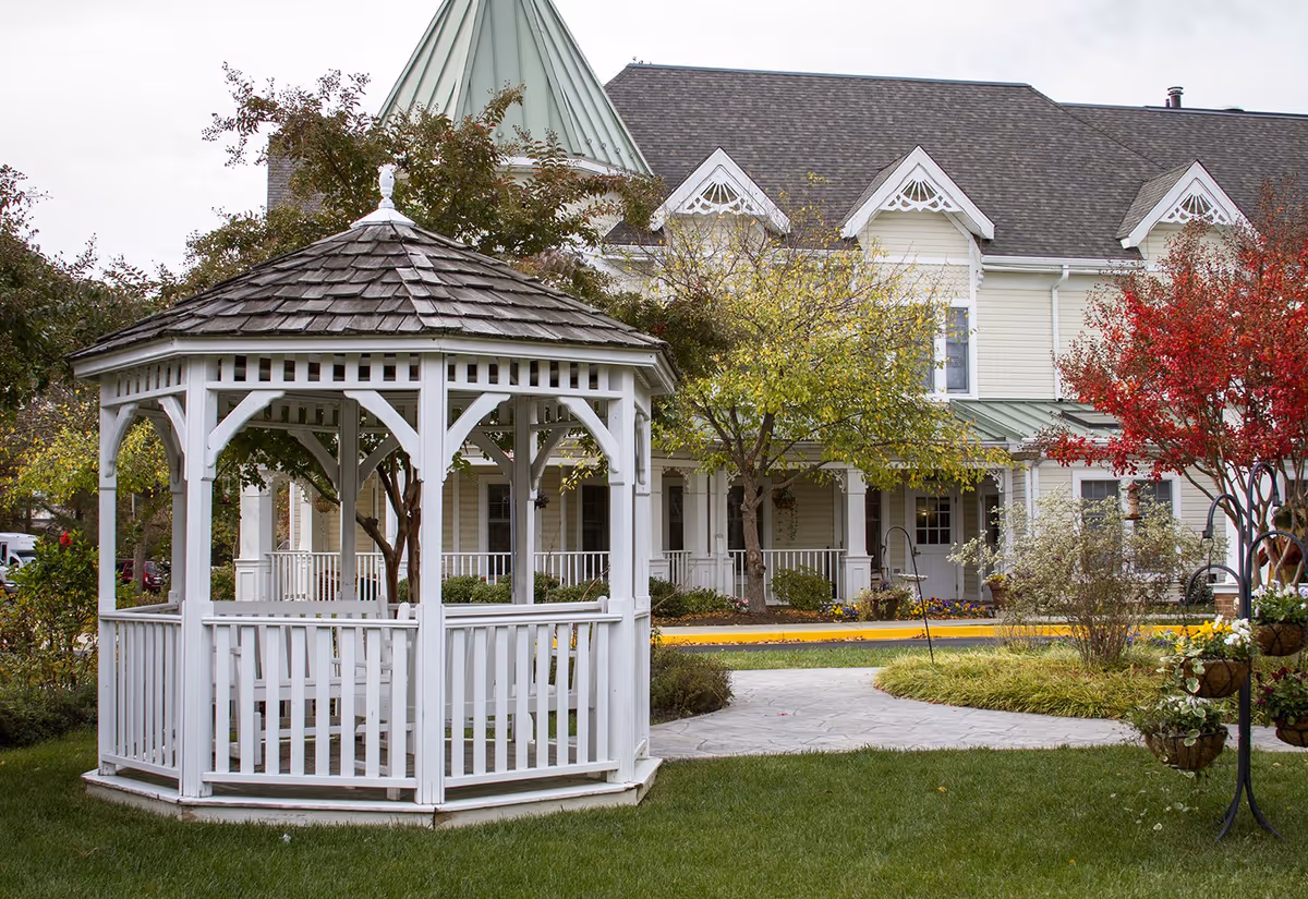 A white wooden gazebo with a shingled roof sits on a green lawn in front of a large, light-colored building with a peaked roof and decorative trim. Trees with green and red leaves surround the gazebo, and a paved walkway curves nearby.