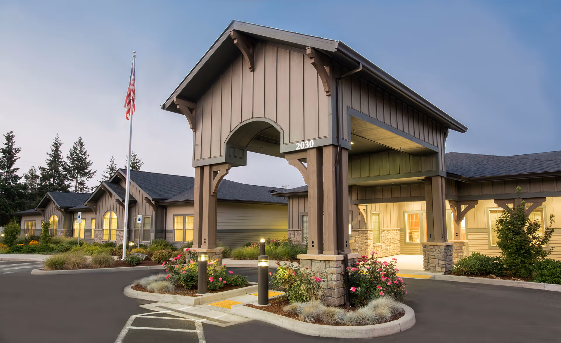 Exterior view of Windsong at Eola Hills facility entrance with a covered drop-off area, landscaped flower beds, and an American flag on a flagpole. The building has a modern design with stone and wood accents, and warm lighting is visible inside.
