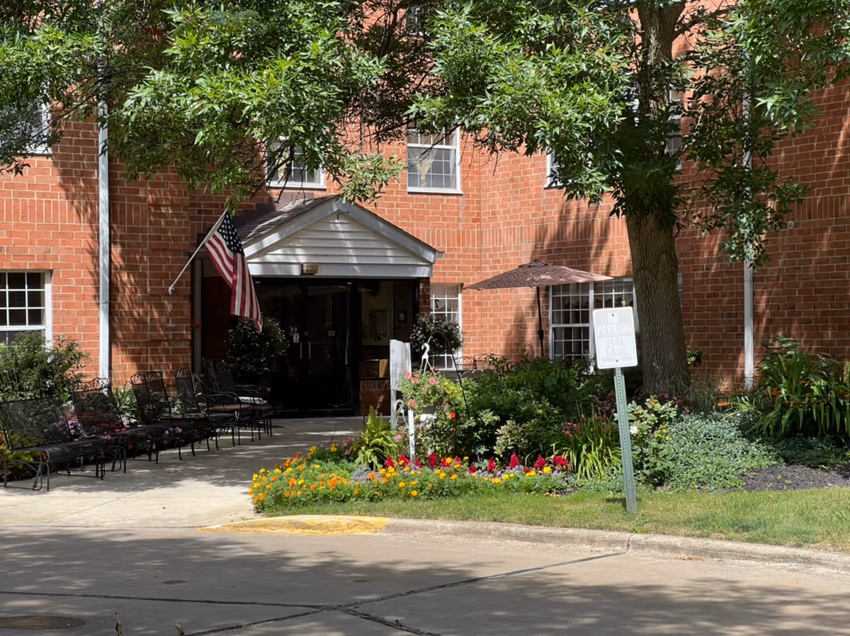Front entrance of a red-brick senior living building with an American flag, benches, and a flower bed.