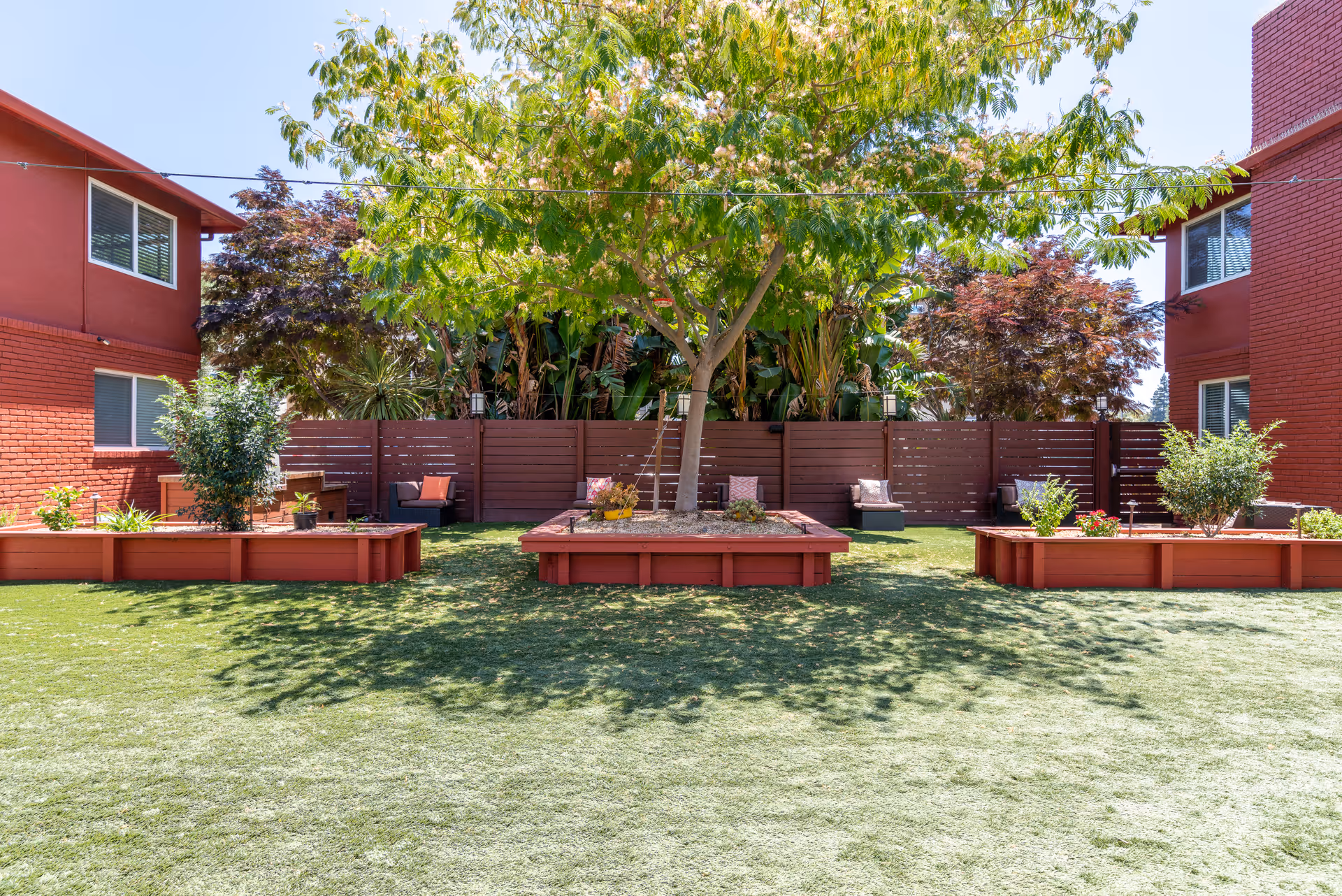 Outdoor courtyard area at Priya Living Santa Clara featuring a large tree in the center surrounded by raised garden beds with plants. There are two red brick buildings on either side and a wooden fence in the background with some outdoor seating.