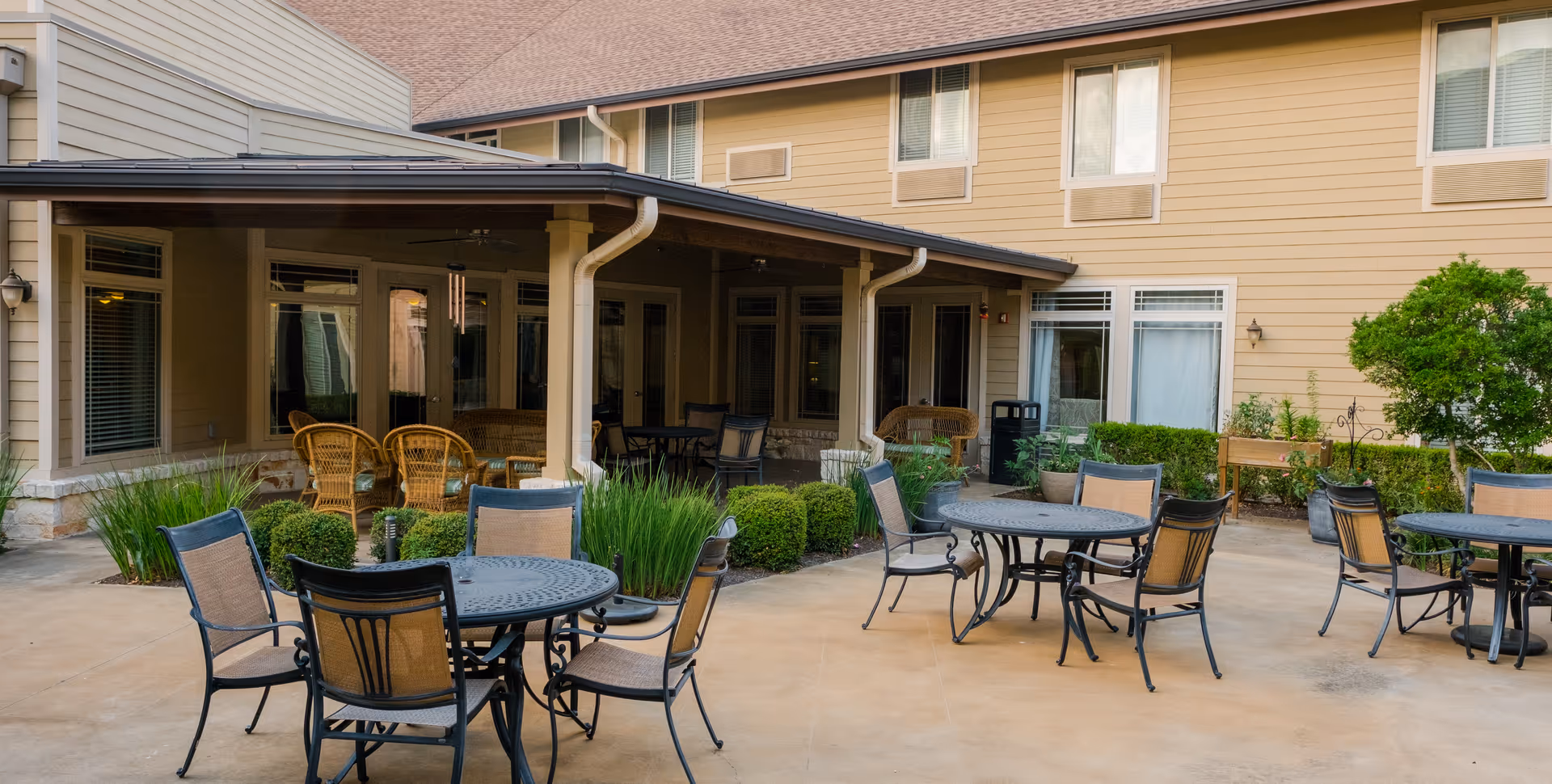 Outdoor patio area at The Pavilion at Great Hills featuring multiple round metal tables with chairs arranged around them, surrounded by greenery and bushes. The patio is adjacent to a beige building with several windows and a covered seating area with wicker chairs.