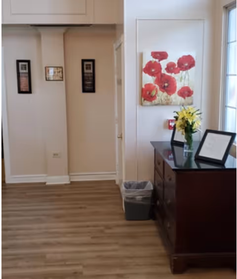 A bright interior hallway with wood flooring, a dark wood dresser topped with a vase of yellow flowers and framed items, red poppy wall art, and a small trash bin.
