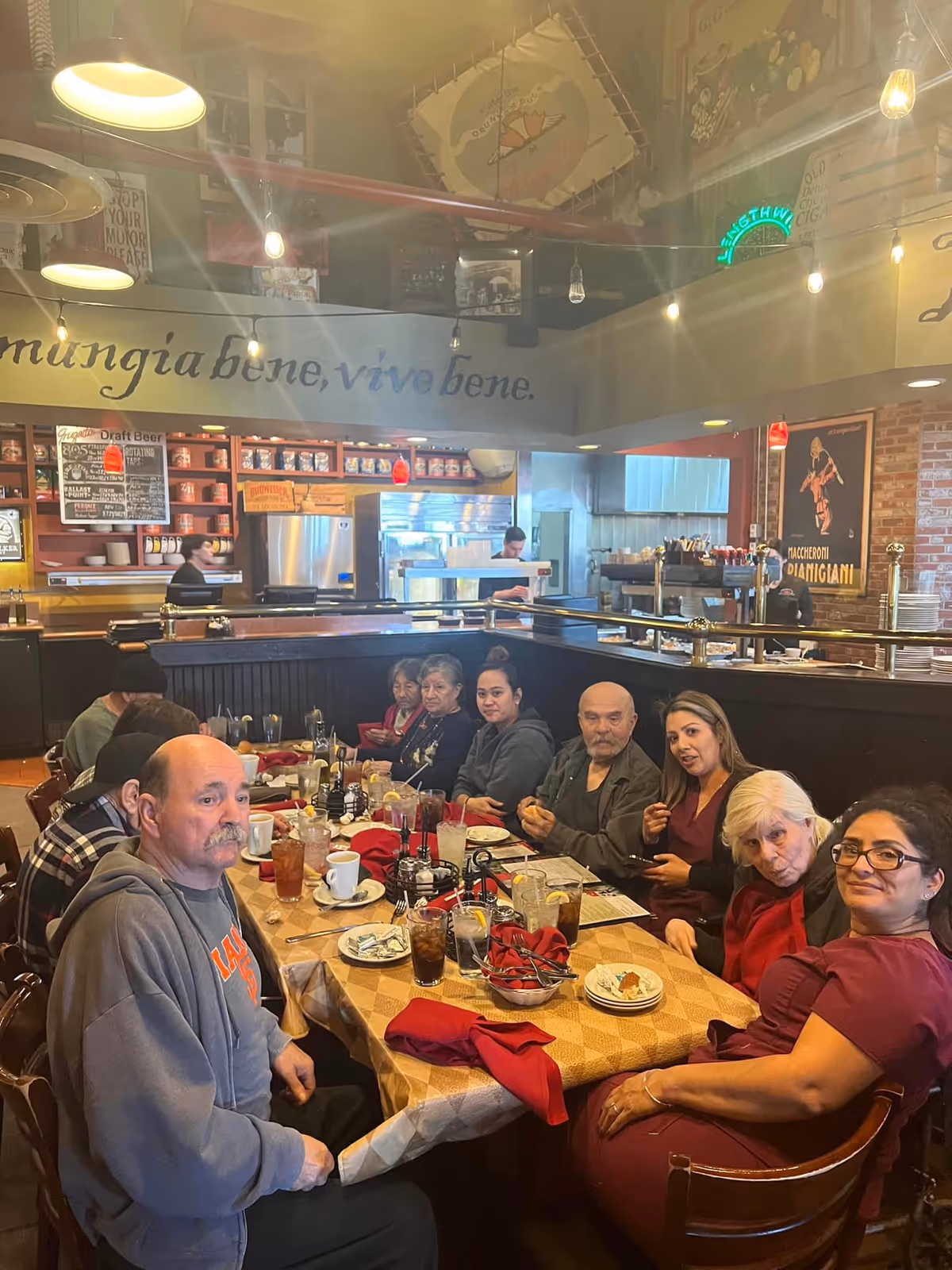 A group of people seated around a large table with plates, drinks, and red napkins in a restaurant-style dining area.