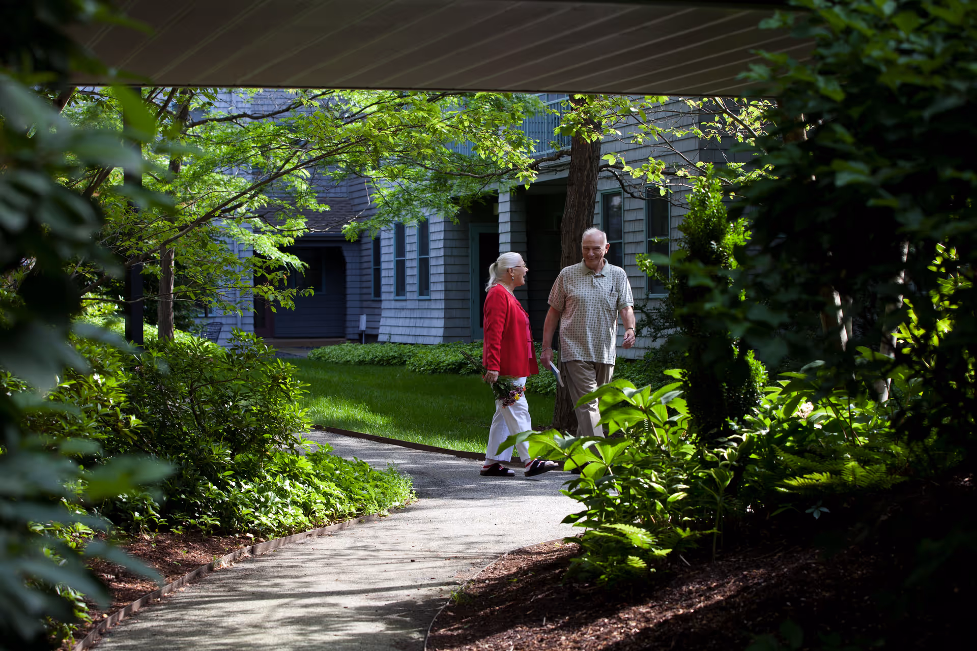 An elderly man and woman walking and talking on a curved garden path surrounded by lush green plants and trees, with a building in the background.