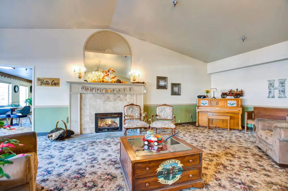 A cozy senior living room with a floral carpet, two floral upholstered chairs, a wooden coffee table with a small red wagon decoration, a fireplace with a 'Happy Fall' banner above it, a piano with decorative items on top, and a sign indicating 'The Bistro Cafe' through an open doorway.