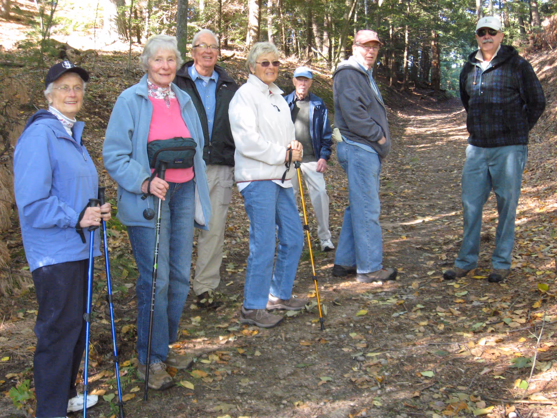 A group of seven elderly people standing on a wooded trail, dressed in casual outdoor clothing and holding walking sticks, surrounded by trees and fallen leaves.