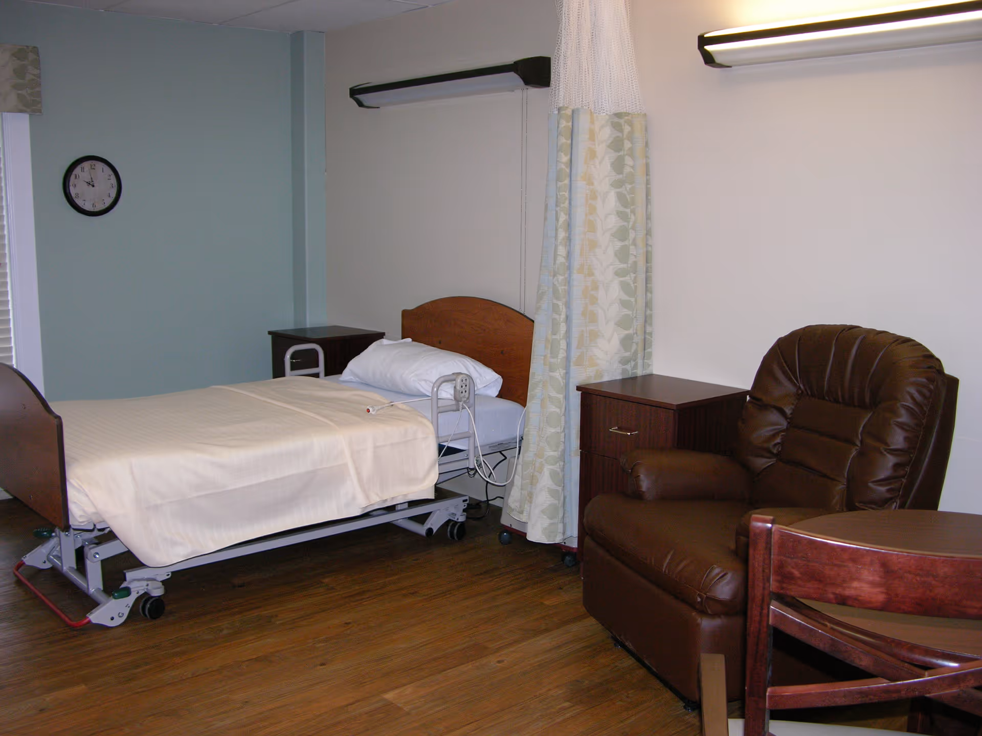 A senior living facility room with a hospital-style bed covered with a beige sheet, a brown leather recliner chair, a wooden side table, a wooden chair, and a privacy curtain. The room has light-colored walls and wooden flooring, with a clock on the wall showing the time as 3:15.