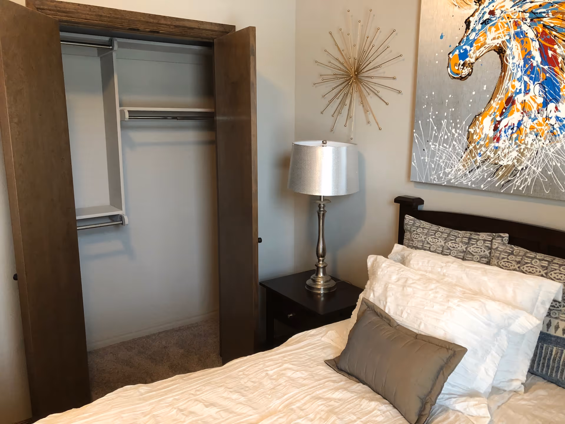 A bedroom featuring a bed with white and gray pillows, a dark wooden headboard, a nightstand with a silver lamp, an open closet with wooden bi-fold doors, and a colorful horse painting on the wall above the bed.