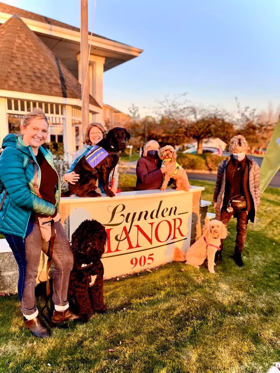 Four women standing and sitting around a sign that reads 'Lynden Manor 905' outside a building, with four dogs on leashes or sitting on the sign. The women are smiling and wearing jackets, and the scene is set on a grassy area with trees and a clear sky in the background.