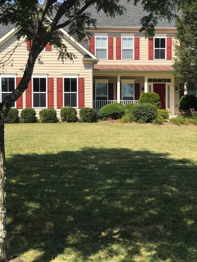 Front exterior view of a two-story house with beige siding and red shutters. The house has a covered porch with white columns, a red front door, and well-maintained bushes and lawn in the foreground. A tree is partially visible on the left side casting shadows on the grass.