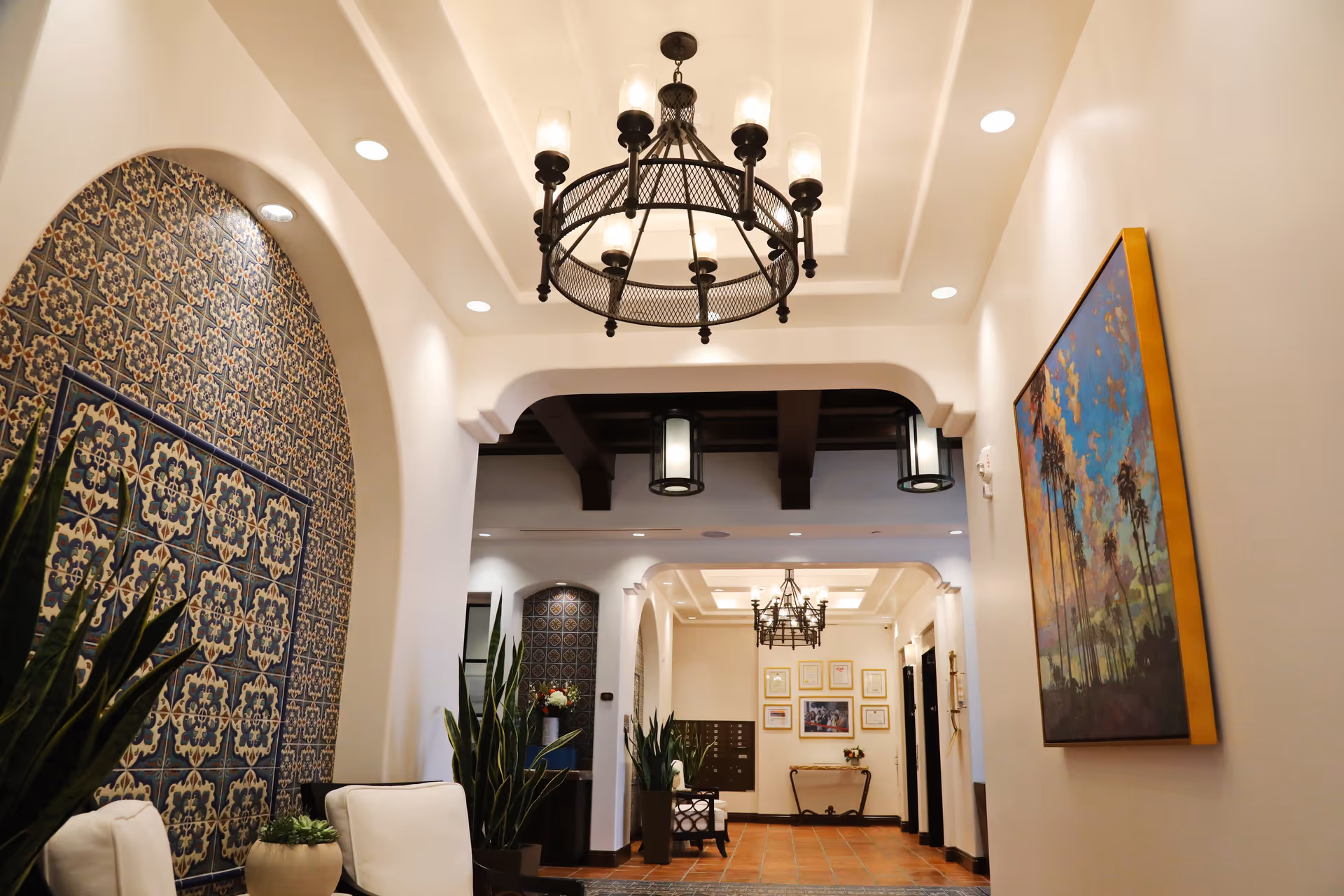 Interior hallway of a senior living facility with white walls, decorative blue and beige tiled arches, potted plants, cushioned chairs, a large chandelier hanging from a recessed ceiling, and a colorful framed painting of palm trees on the right wall.