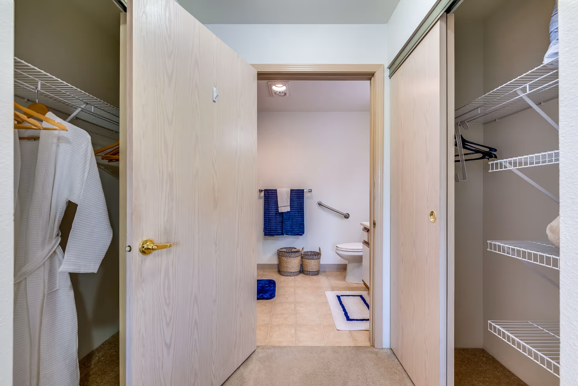 View through an open door into a bathroom with beige tiled floor, a toilet, two woven baskets, and blue and white towels hanging on a rack. On either side of the door are closets with wire shelving and hangers, one closet containing a white robe.