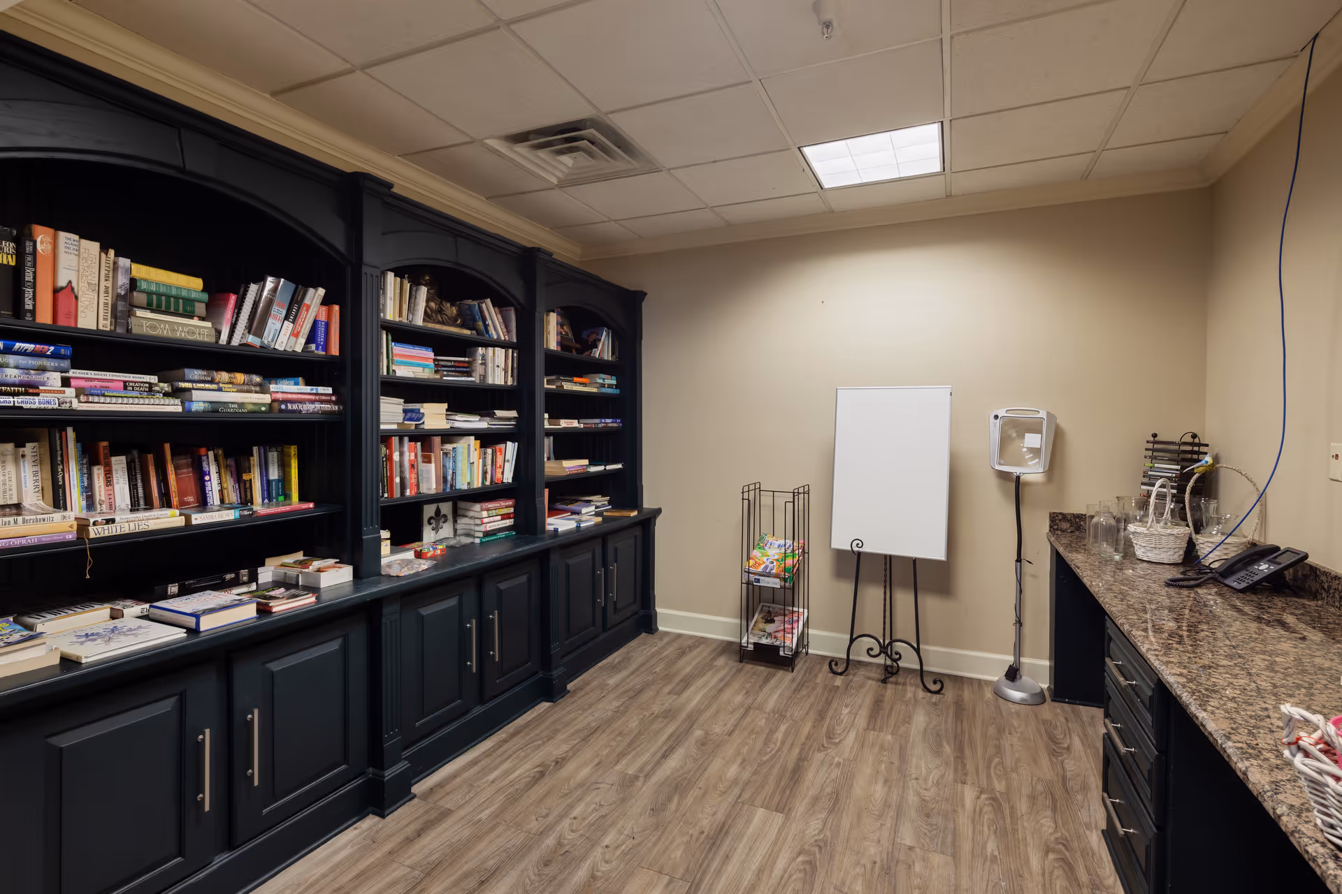 Interior room with dark wood bookshelves filled with books on the left side, a wooden floor, a blank white easel, a small metal rack with magazines, and a countertop with a phone, glasses, and baskets on the right side.