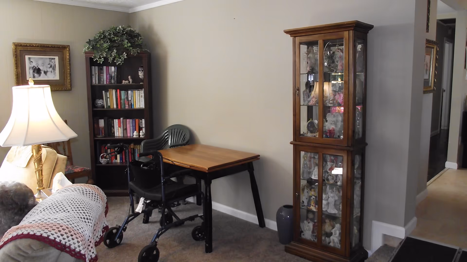 A cozy living room area with a beige couch covered by a crocheted blanket, a wooden bookshelf filled with books and decorative items, a wooden table with a black walker in front of it, a green plastic chair, a tall wooden display cabinet filled with porcelain figurines, a table lamp with a white shade, and framed pictures on the walls.