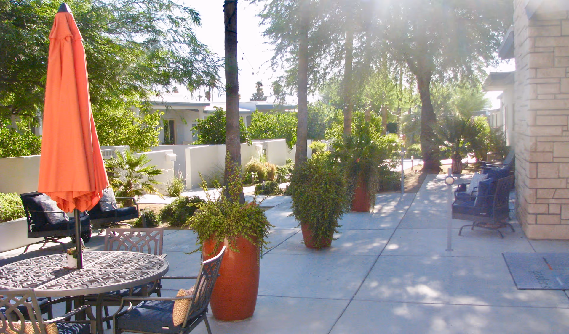 Outdoor patio area with a round metal table and chairs, an orange closed umbrella, large potted plants, and benches along a walkway surrounded by trees and greenery.