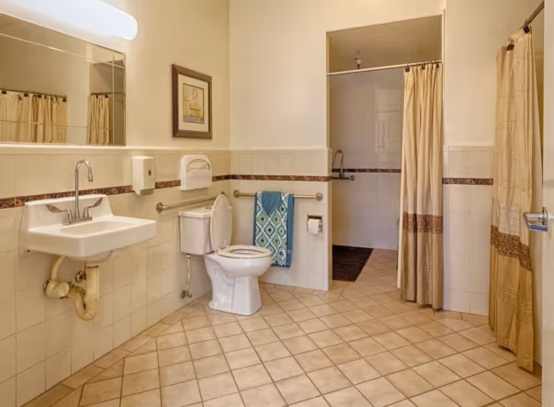 A clean and spacious bathroom with beige tiled floor and walls. It features a white sink with a silver faucet, a toilet with the lid open, a blue patterned towel hanging on a grab bar, and a toilet paper holder. There are two shower areas with beige curtains, one visible in the background and another partially visible on the right. A framed picture hangs on the wall above the toilet, and a large mirror is mounted above the sink.
