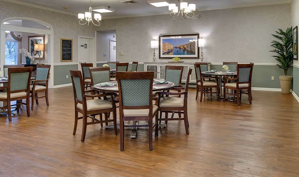 Dining room with round tables and wooden chairs on a hardwood floor in a senior living facility.