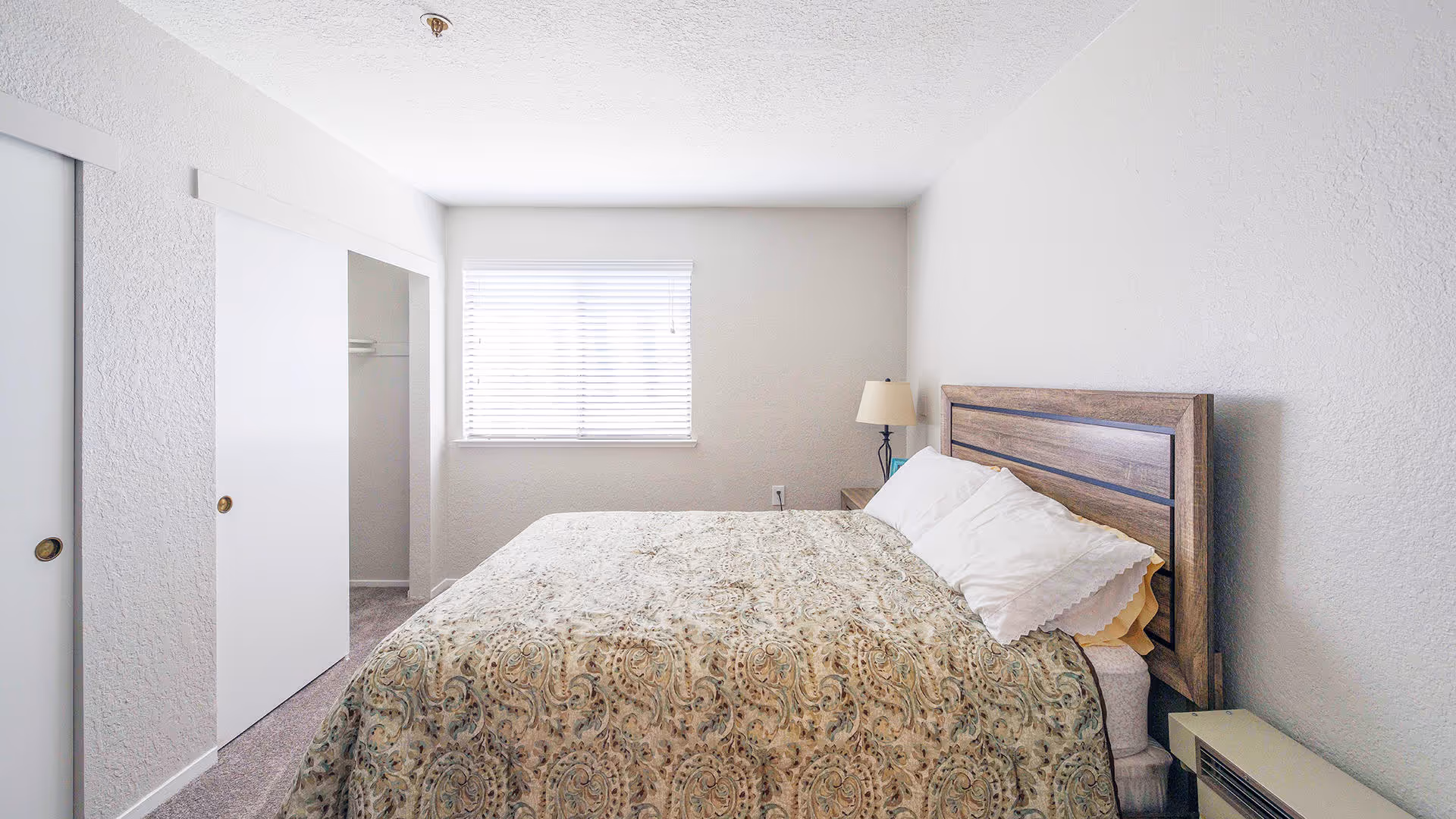 A bright bedroom with a large bed covered in a patterned bedspread and white pillows. There is a wooden headboard, a bedside table with a lamp, a window with blinds, and a closet with sliding doors.