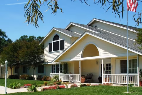 Exterior view of Oakwood Meadows Assisted Living building with a well-maintained lawn, shrubs, and trees. The building features light-colored siding, a covered porch with seating, and an American flag on a flagpole in the front yard under a clear blue sky.