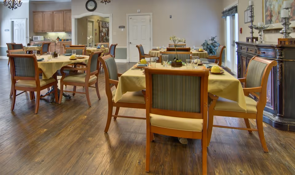 A bright dining room with several tables set with yellow tablecloths and wooden chairs.