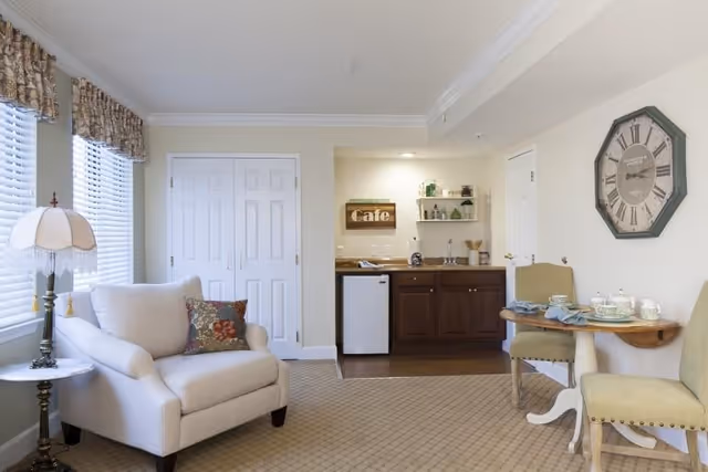 A cozy living area with a beige armchair next to a window with blinds and floral valances. A small round table with a vintage lamp is beside the armchair. In the background, there is a kitchenette with dark wood cabinets, a small refrigerator, and a sign that says 'Cafe'. To the right, there is a small wooden dining table set with two green upholstered chairs and tea set. A large wall clock hangs above the dining area.