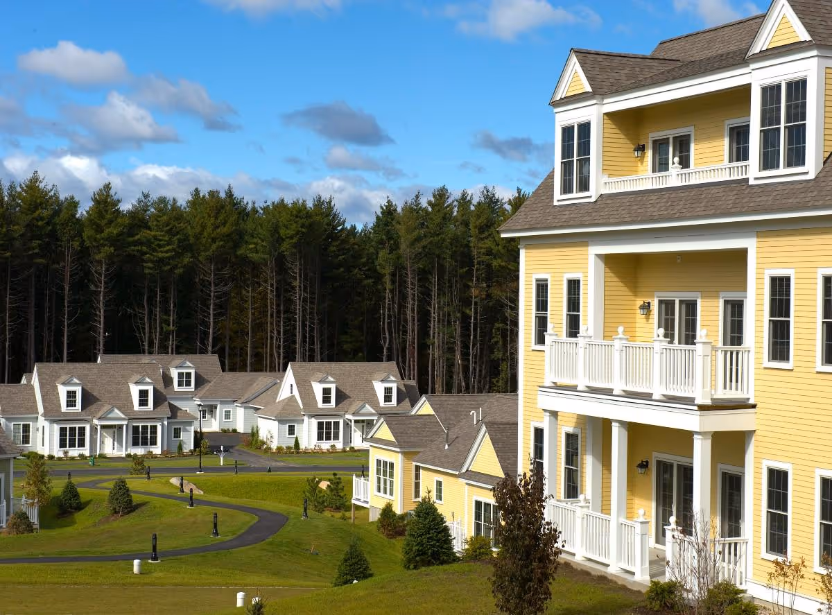 Exterior view of a senior living facility with multiple yellow and white residential-style buildings surrounded by green lawns and trees under a blue sky with some clouds.