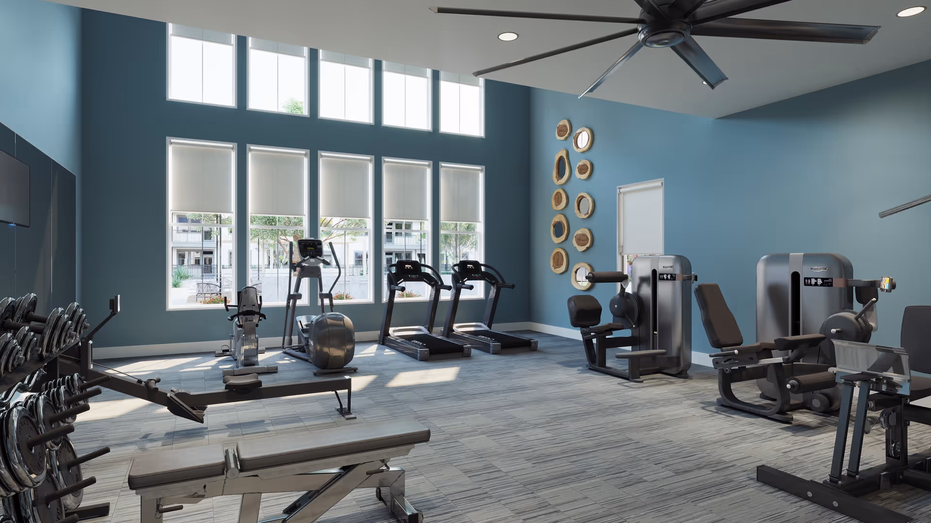 A bright and spacious fitness room with large windows letting in natural light. The room features various exercise equipment including treadmills, weight machines, a rowing machine, and free weights. The walls are painted blue and decorated with circular wooden wall art. A large ceiling fan is mounted above.