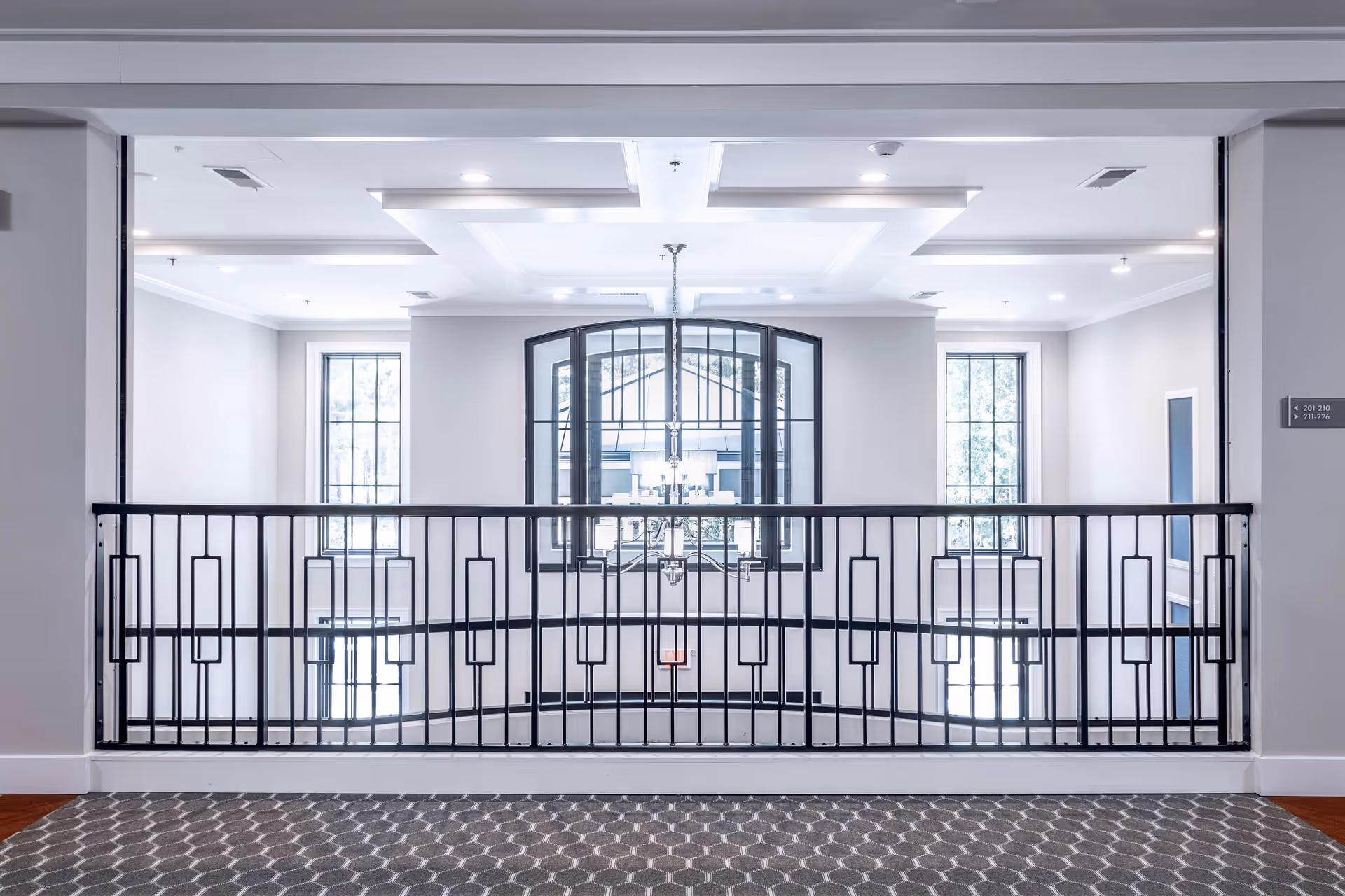 Interior view of a senior living facility hallway with a decorative black metal railing overlooking a lower level. The walls are painted light gray with white trim, and there are two tall windows letting in natural light. A modern chandelier hangs in the center, and the ceiling has recessed lighting and a coffered design. The floor is covered with a patterned gray carpet.