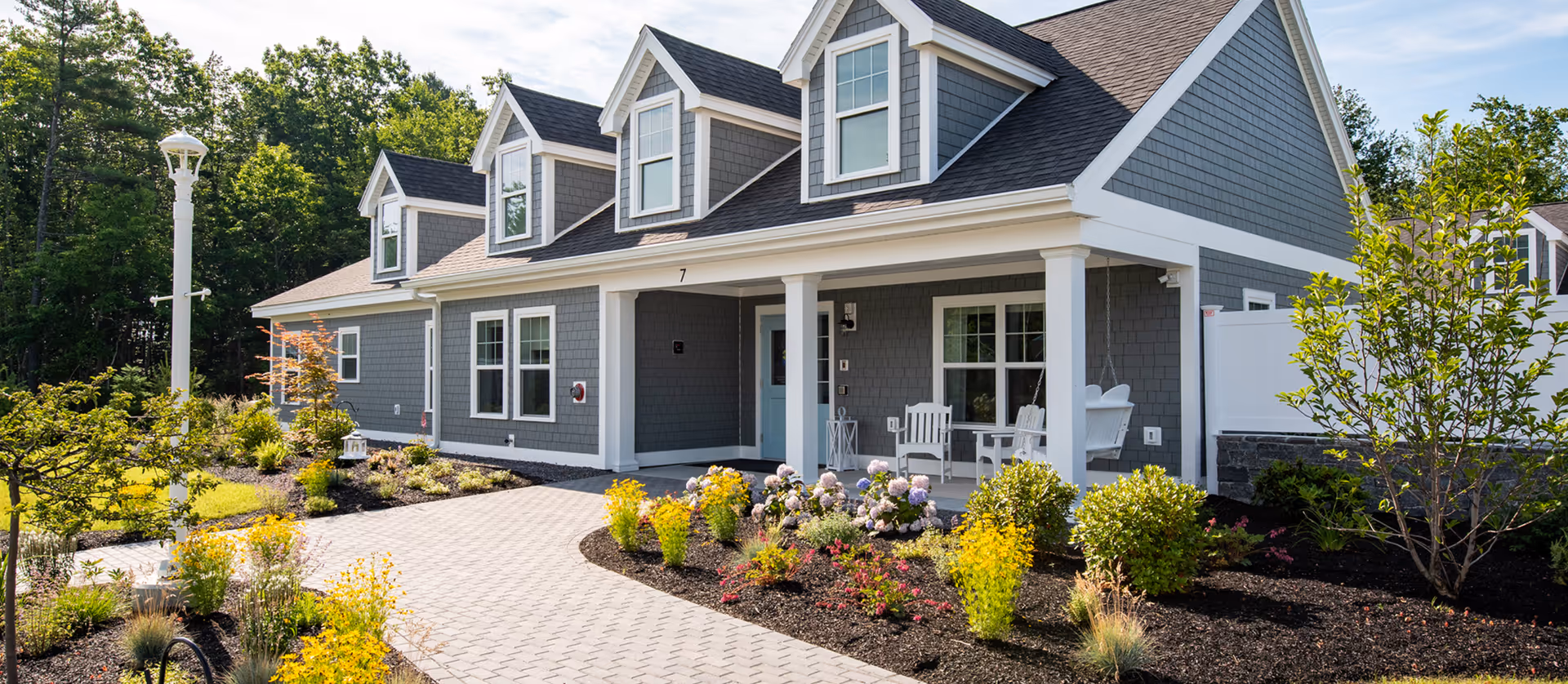 Gray and white single-story building with dormer windows, a covered porch with chairs, a paved walkway and landscaped gardens in front.