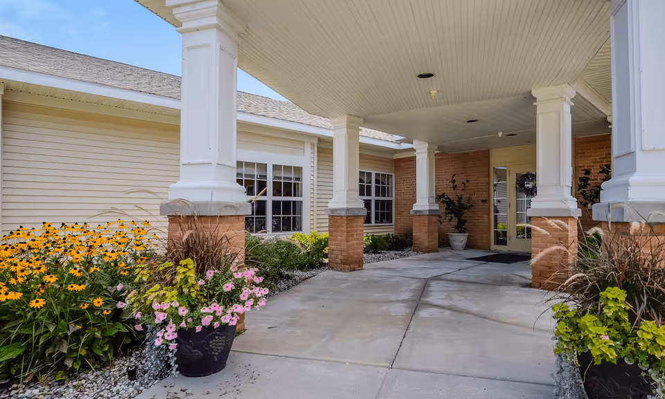 Covered entrance area of a building with white columns and brick bases, surrounded by flower pots with colorful flowers and green plants, and beige siding walls with windows.