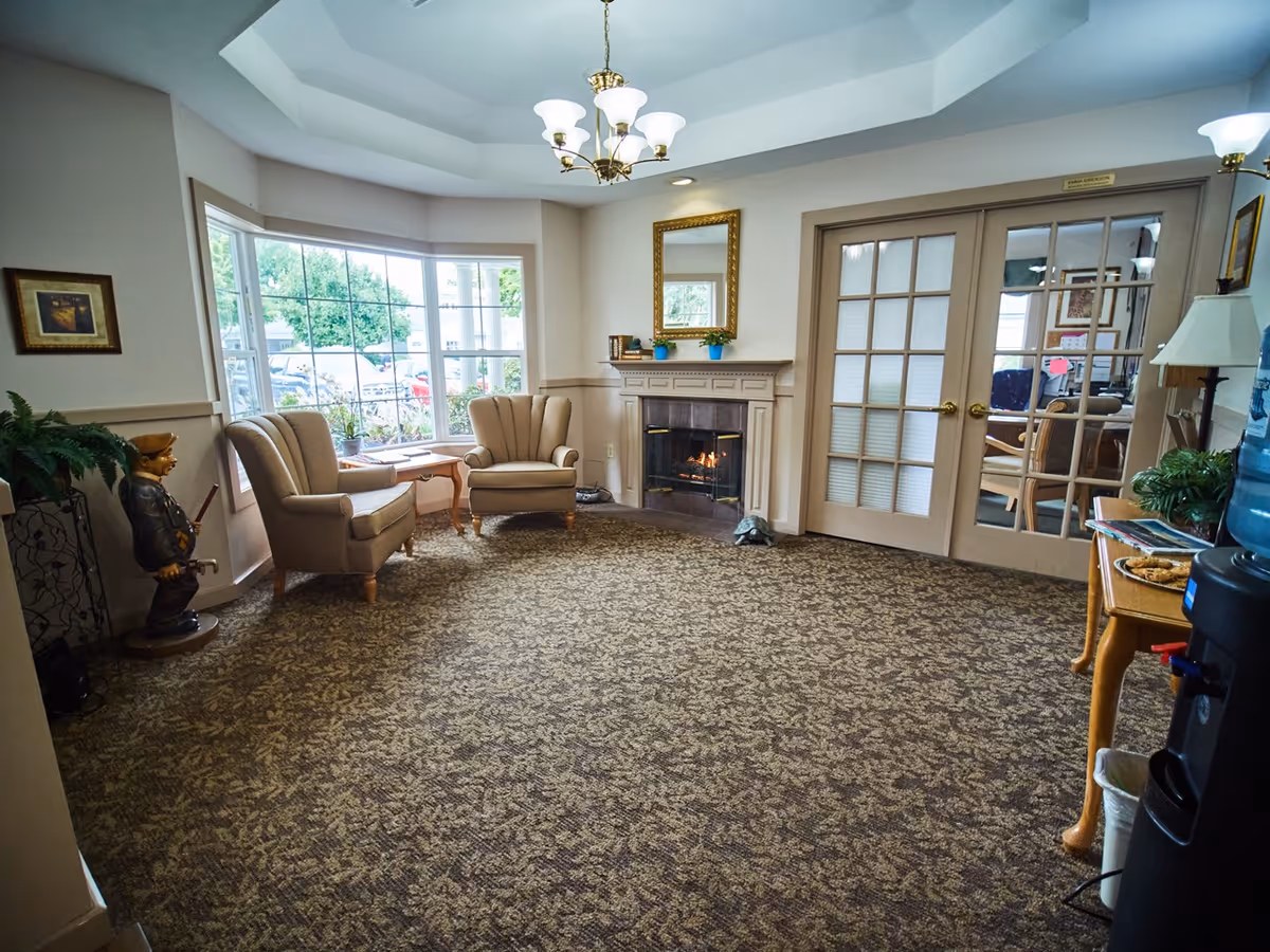 A senior living lounge with two armchairs by a bay window, a lit fireplace, and glass double doors.