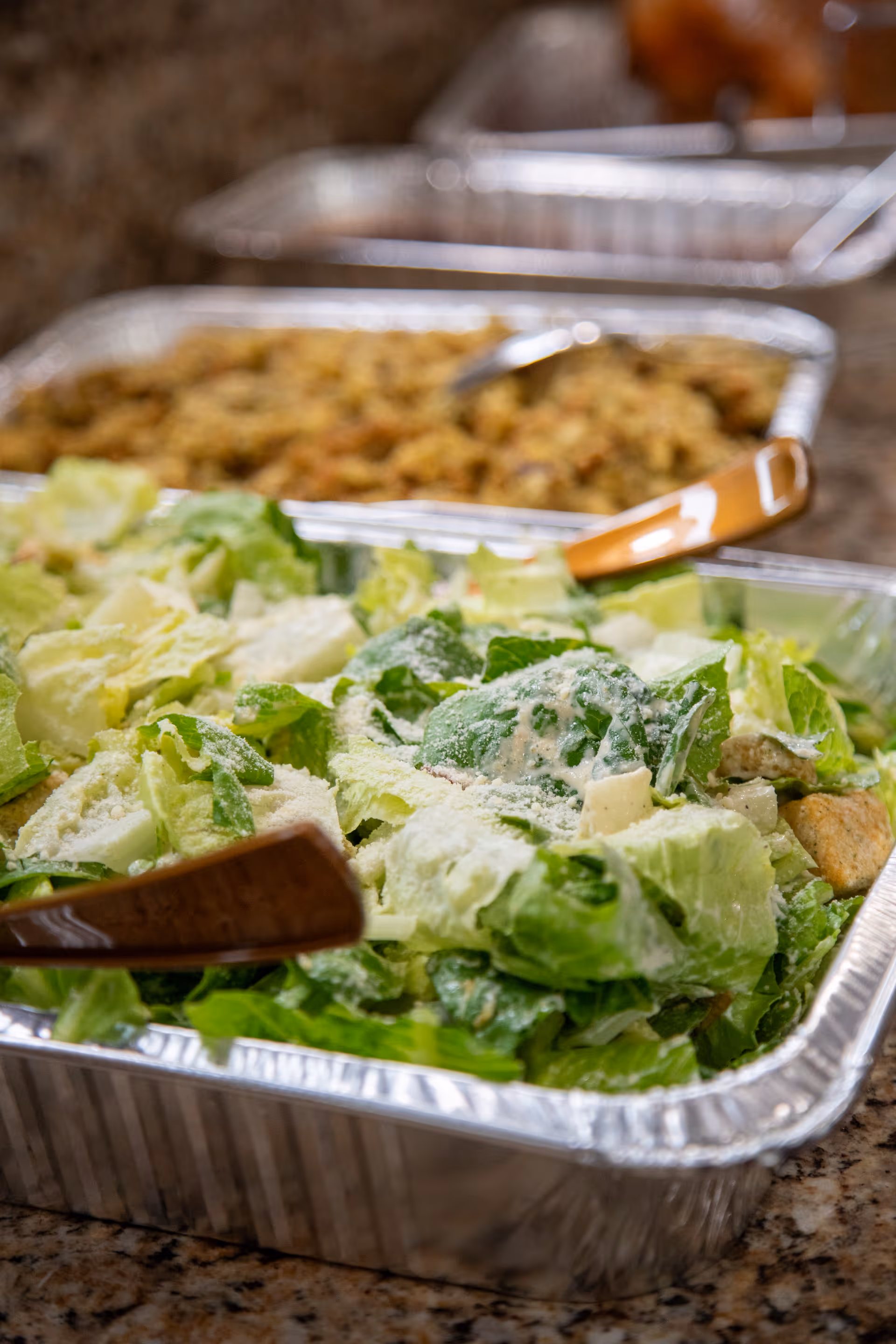 Close-up of a buffet tray filled with Caesar salad and wooden serving utensils, with additional trays of food blurred in the background.