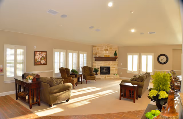A spacious living room with beige walls and carpet, featuring multiple cushioned armchairs and sofas arranged around a stone fireplace. The room is well-lit with natural light coming through several windows with white shutters. There are wooden side tables with decorative plants and a painting on the wall. The ceiling has recessed lighting and built-in speakers.