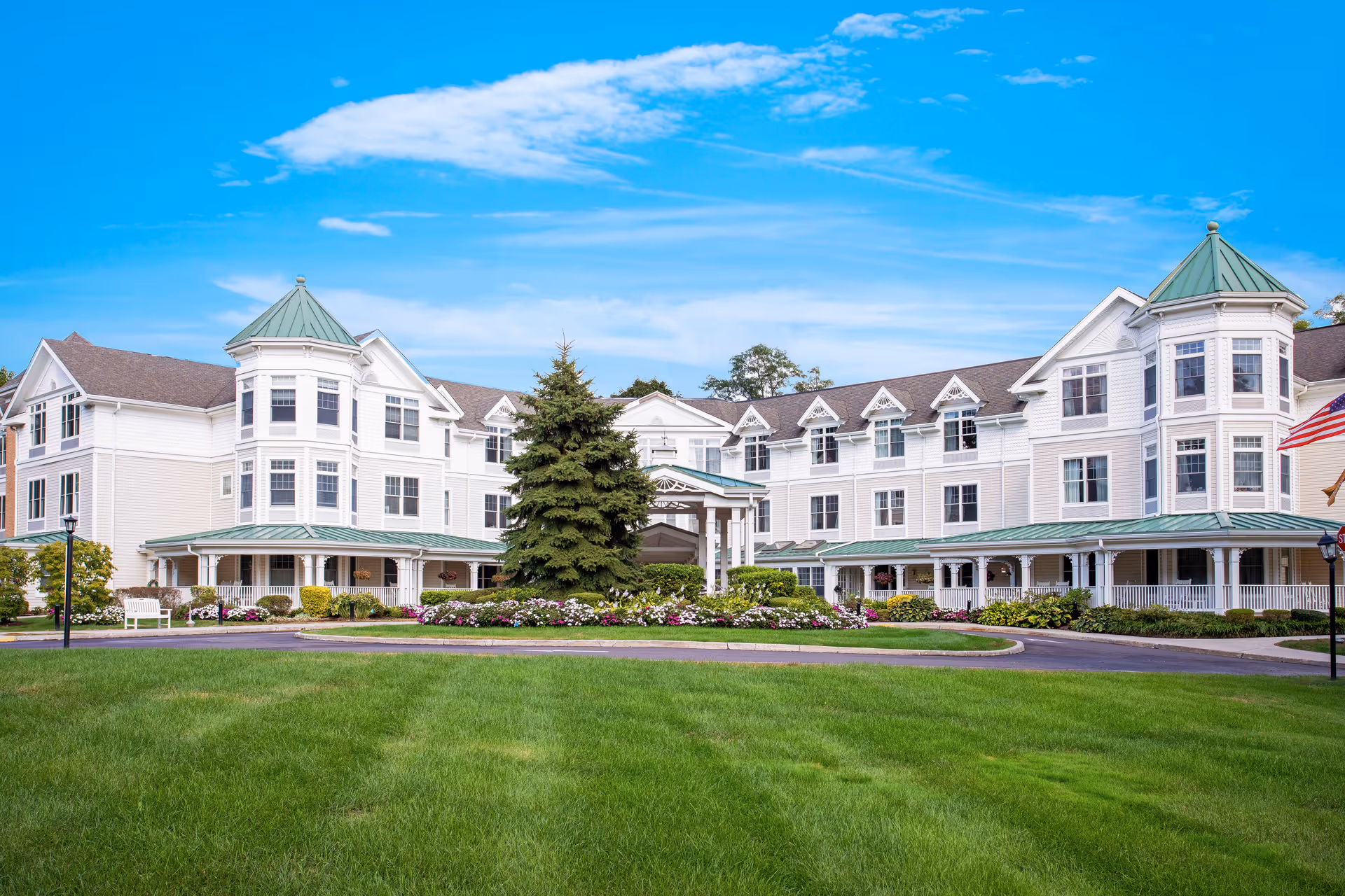 Front exterior view of a large, white senior living facility building with green roofs and multiple windows. The building is surrounded by a well-maintained lawn, flower beds, and a large evergreen tree in the center. The sky is clear and blue with some clouds.