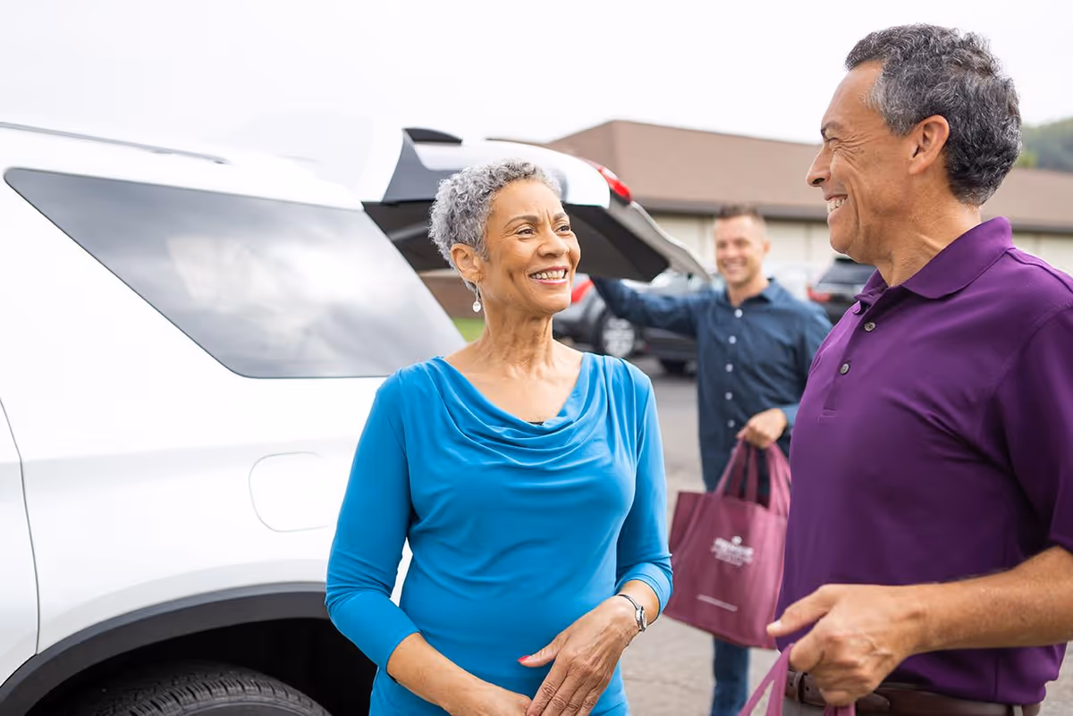 An older woman in a blue top and an older man in a purple polo shirt smile and talk near a white vehicle with its trunk open. In the background, a younger man is holding a maroon tote bag and standing near the vehicle in a parking lot with a building behind them.