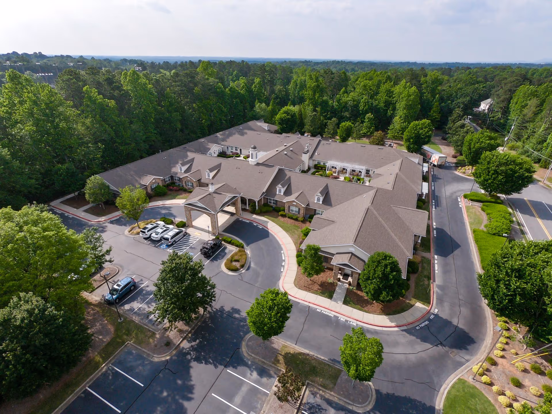 Aerial view of a single-story senior living facility with a porte-cochère, circular driveway, parking lot, and surrounding trees.