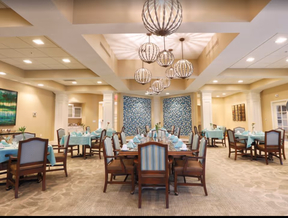 A spacious dining room with several wooden tables and chairs arranged neatly. The tables are covered with light blue tablecloths and set with napkins, glasses, and small flower vases. The ceiling features multiple decorative spherical pendant lights casting patterned shadows. The walls are beige with two large blue patterned panels and some artwork. The room has a carpeted floor and a bright, welcoming atmosphere.