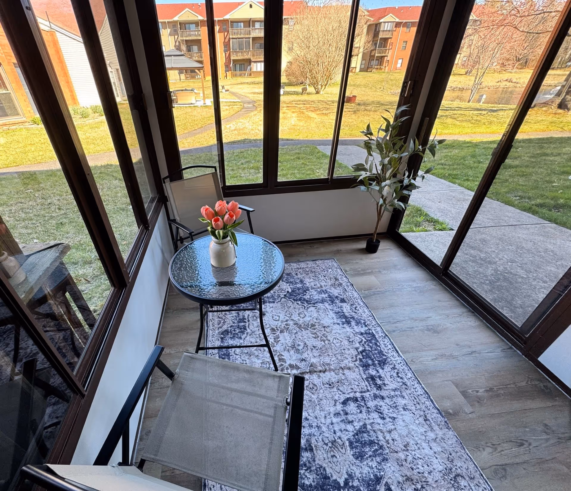 A small enclosed patio area with large windows overlooking a grassy courtyard and apartment buildings. Inside the patio, there is a glass-top round table with a vase of pink tulips, two mesh chairs, a patterned rug on the floor, and a potted plant in the corner.