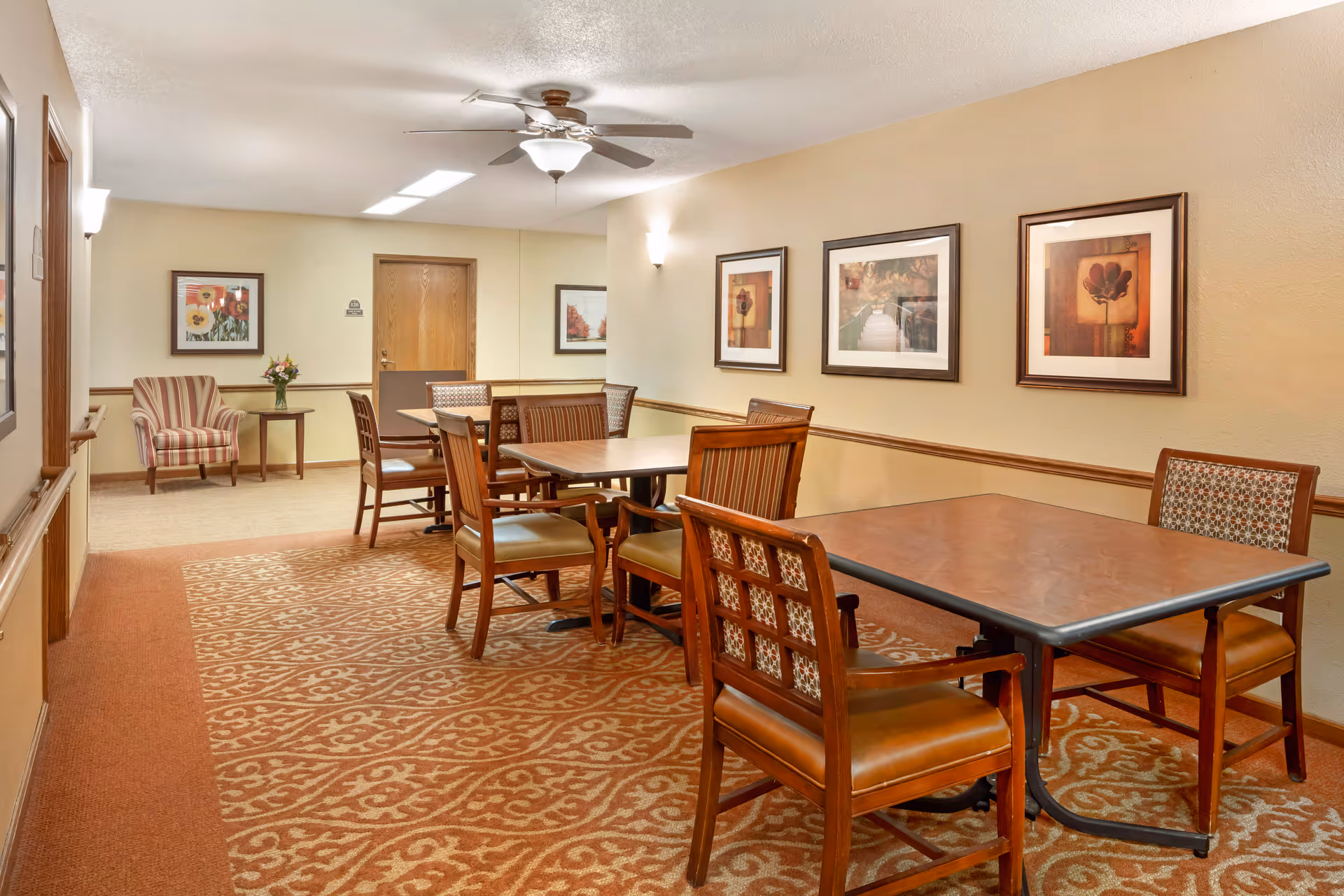 Interior view of a common area in a senior living facility with multiple wooden tables and chairs arranged on a patterned carpet. The walls are decorated with framed artwork and there is a ceiling fan with a light fixture. A striped armchair and a small table with a flower vase are visible in the background near a wooden door.
