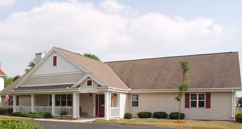 Single-story beige building with a covered front porch, small lawn and walkway under a partly cloudy sky.