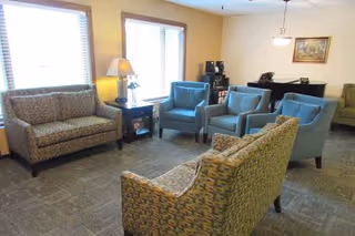 A cozy living room area with patterned sofas and blue armchairs arranged around a small table with a lamp. There are two large windows with blinds letting in natural light, and a piano with a framed picture hanging above it in the background.