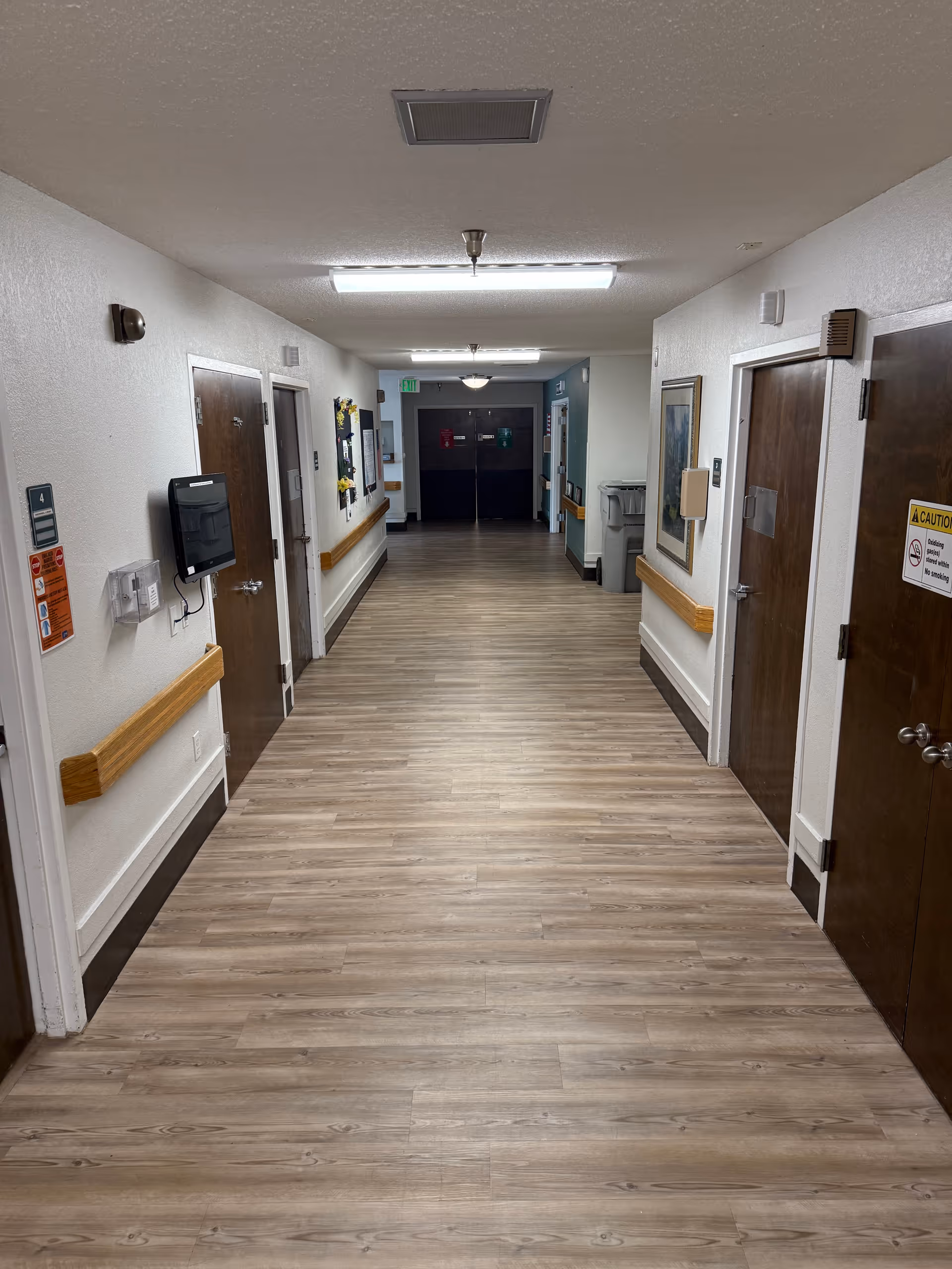 Long wood-floored interior hallway with doors on both sides, handrails along the walls, and overhead lighting.