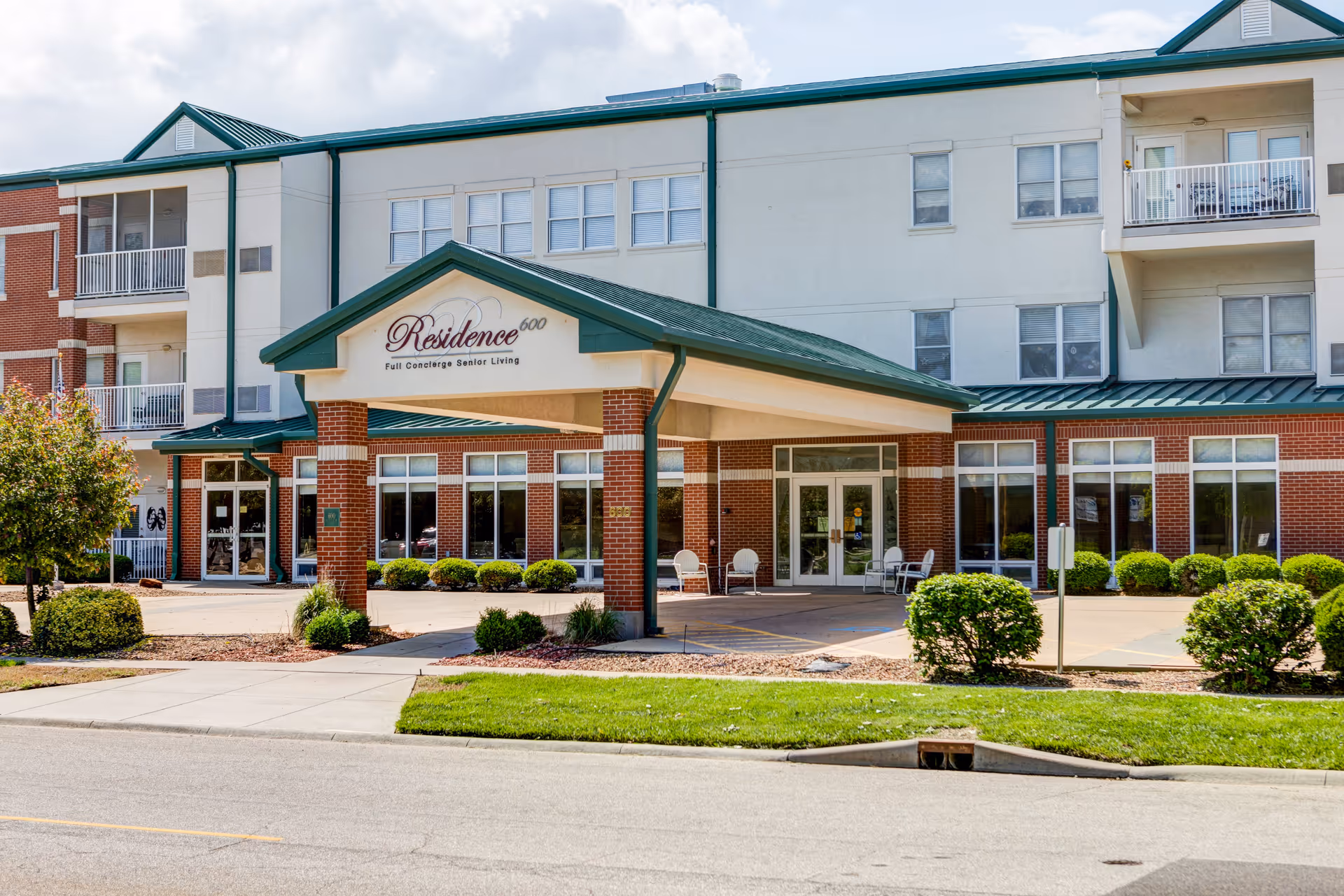 Exterior view of Residence 600, a senior living facility with a covered entrance supported by brick columns, green roof, and multiple windows. There are bushes and a small tree in front, along with a sidewalk and a street in the foreground.