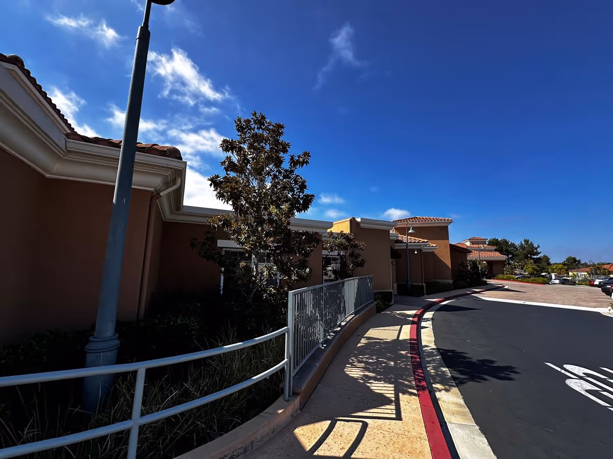 Exterior of a single-story building with a curved driveway, sidewalk railing, lamp post and trees under a bright blue sky.