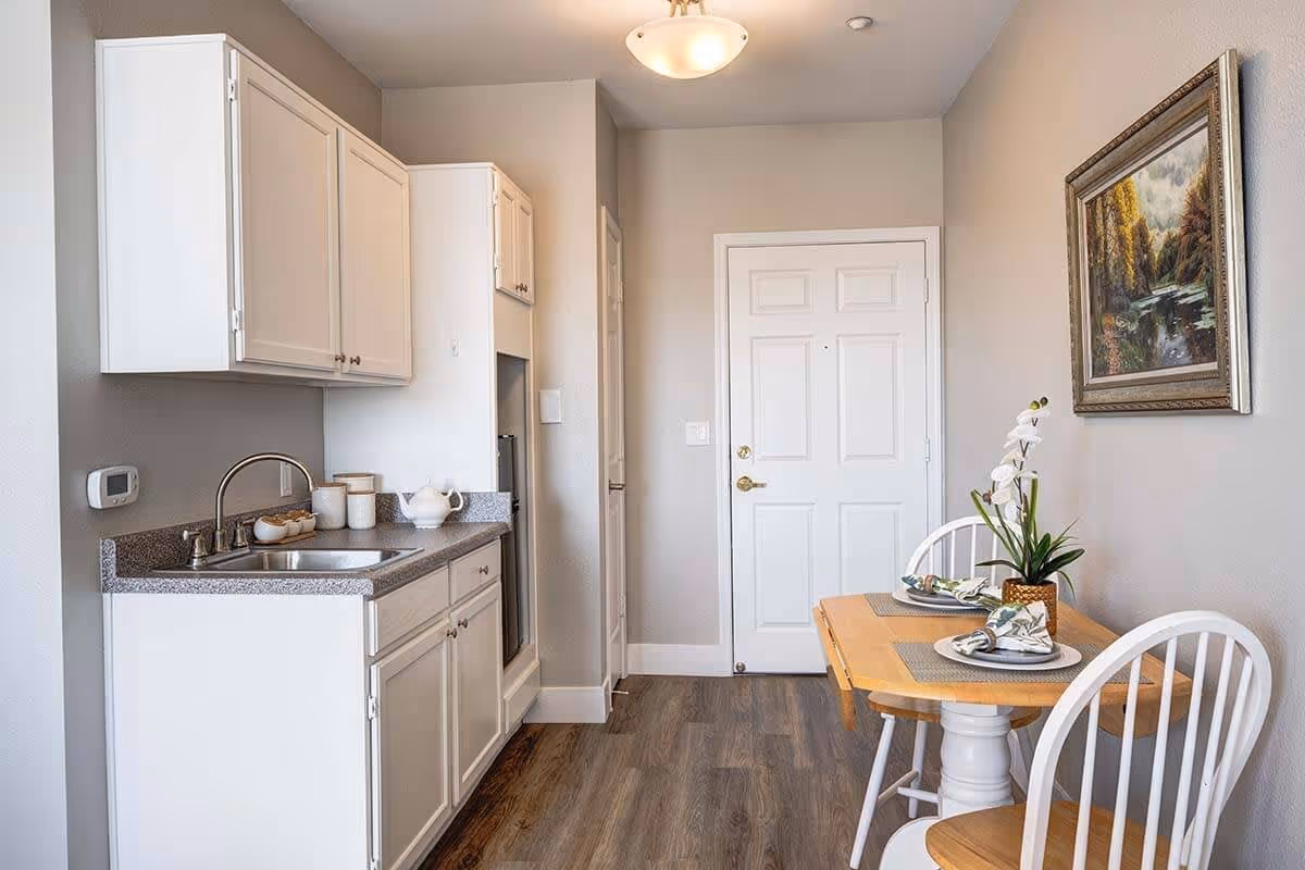 A small kitchen and dining area in an assisted living facility. The kitchen features white cabinets, a stainless steel sink, and a countertop with some kitchen items. Adjacent to the kitchen is a wooden dining table set for two with white chairs, a potted plant centerpiece, and a framed landscape painting on the wall. The floor is wood-style laminate, and a white door is visible at the end of the room.
