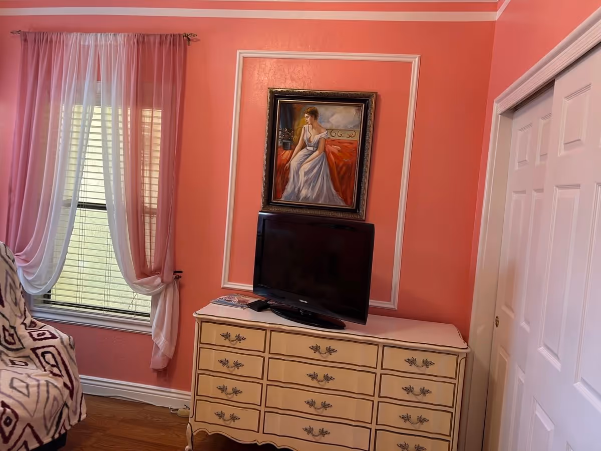 A cozy room with coral pink walls featuring a window with sheer white and pink curtains, a cream-colored dresser with ornate handles, a flat-screen TV on top of the dresser, and a framed painting of a woman in a white dress hanging above the TV. There is also a patterned armchair partially visible on the left and white sliding closet doors on the right.