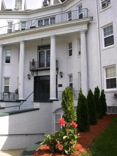 Front exterior view of a white multi-story building with large white columns, black railings, and several windows. There is a small garden area with green shrubs and red flowers in front of the building.